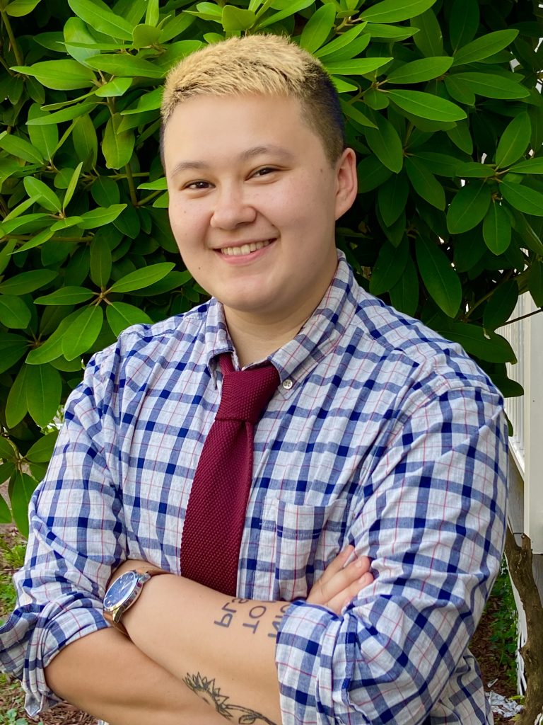 A photo of MG from the waist up, wearing a blue and white plaid shirt with a red tie. The top of their hair is bleached. They are standing in front of a large green plant.