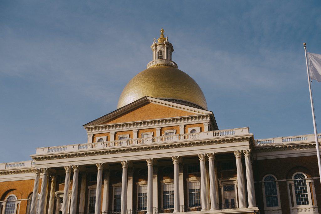 A image of the Massachusetts State house. Tilted upward, it emphasizes the golden dome and white columns and sits against a light blue sky.